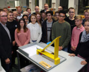 Eine Gruppe von Menschen, darunter Jugendliche und Erwachsene, versammelt sich um einen Tisch mit einem gelben Modellbauwerk an der Hochschule Coburg. Sie befinden sich in einem Raum mit verschiedenen Geräten und scheinen in eine lebhafte Diskussion oder Präsentation vertieft zu sein.