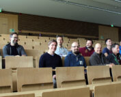 Eine Gruppe von zwölf Personen sitzt in den Reihen eines leeren Hörsaals der Hochschule Coburg. Sie lächeln und tragen legere Kleidung, umgeben von Holzsitzen und -vertäfelungen und einer gut beleuchteten Atmosphäre.