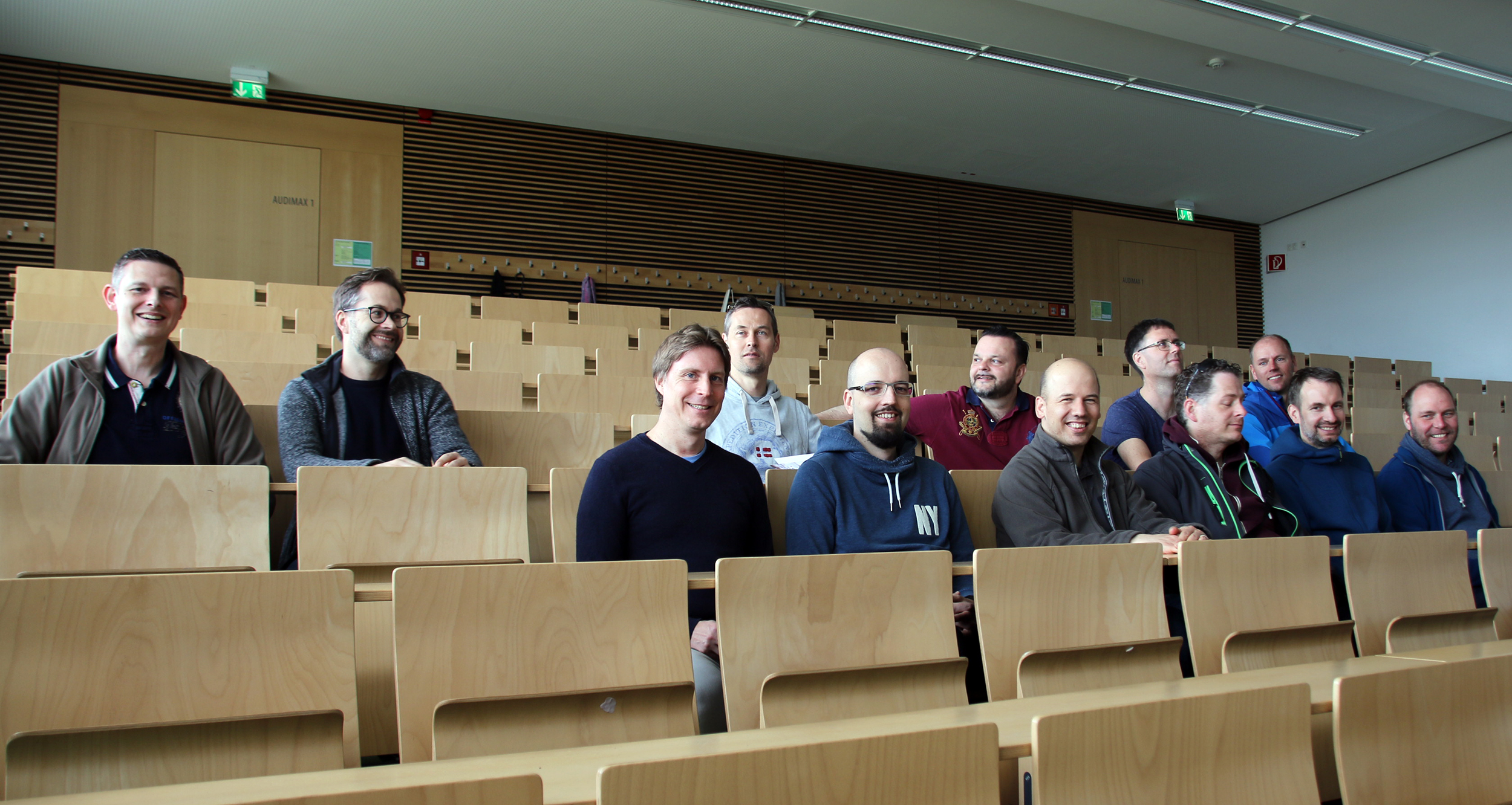 Eine Gruppe von zwölf Personen sitzt in den Reihen eines leeren Hörsaals der Hochschule Coburg. Sie lächeln und tragen legere Kleidung, umgeben von Holzsitzen und -vertäfelungen und einer gut beleuchteten Atmosphäre.