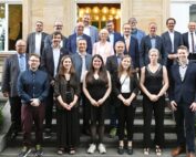 Eine Gruppe von Menschen, Männer und Frauen, steht auf der Treppe vor einem Gebäude der Hochschule Coburg. Einige sind formell gekleidet und tragen Anzüge, andere sind eher leger gekleidet. Sie stehen in mehreren Reihen, lächeln und posieren für das Foto.