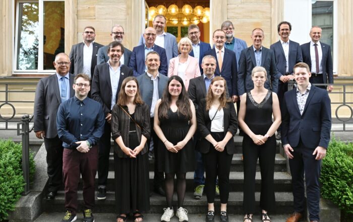 Eine Gruppe von Menschen, Männer und Frauen, steht auf der Treppe vor einem Gebäude der Hochschule Coburg. Einige sind formell gekleidet und tragen Anzüge, andere sind eher leger gekleidet. Sie stehen in mehreren Reihen, lächeln und posieren für das Foto.