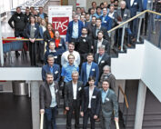 Eine große Gruppe von Menschen, überwiegend Männer in Business-Kleidung, steht auf einer Treppe in einem modernen Gebäude. Sie posieren für ein Gruppenfoto, hinter ihnen ein Banner mit der Abkürzung "TAS". Tische und Stühle daneben deuten auf eine Veranstaltung der Hochschule Coburg hin.