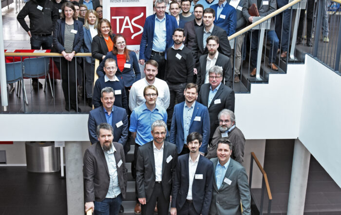 Eine große Gruppe von Menschen, überwiegend Männer in Business-Kleidung, steht auf einer Treppe in einem modernen Gebäude. Sie posieren für ein Gruppenfoto, hinter ihnen ein Banner mit der Abkürzung "TAS". Tische und Stühle daneben deuten auf eine Veranstaltung der Hochschule Coburg hin.
