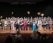 Eine große Gruppe von Menschen der Hochschule Coburg posiert auf einer Bühne und hält Zertifikate und Auszeichnungen in den Händen. Sie tragen formelle Kleidung. Der Hintergrund ist schwarz mit dekorativen goldenen Kronen, während die Zuschauer im Vordergrund den Moment mit ihren Handys festhalten.