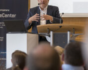 Ein Mann im Anzug steht an einem Podium und hält einen Vortrag. Er vertritt die Hochschule Coburg. Während er spricht, gestikuliert er mit den Händen. Im Hintergrund sind eine Projektionsfläche und ein Banner zu sehen, im Vordergrund sitzt ein aufmerksames Publikum.