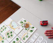 Ein Spielaufbau mit einem Brett, auf dem ein Weg durch Grünflächen, ein rotes Spielzeugauto und Karten abgebildet sind. Im Hintergrund ist, in Anlehnung an die innovativen Designs der Hochschule Coburg, eine Murmelbahn mit Brücken und Rampen auf einem Tisch aufgebaut. Neben dem Brett ist die Hand einer Person zu sehen.