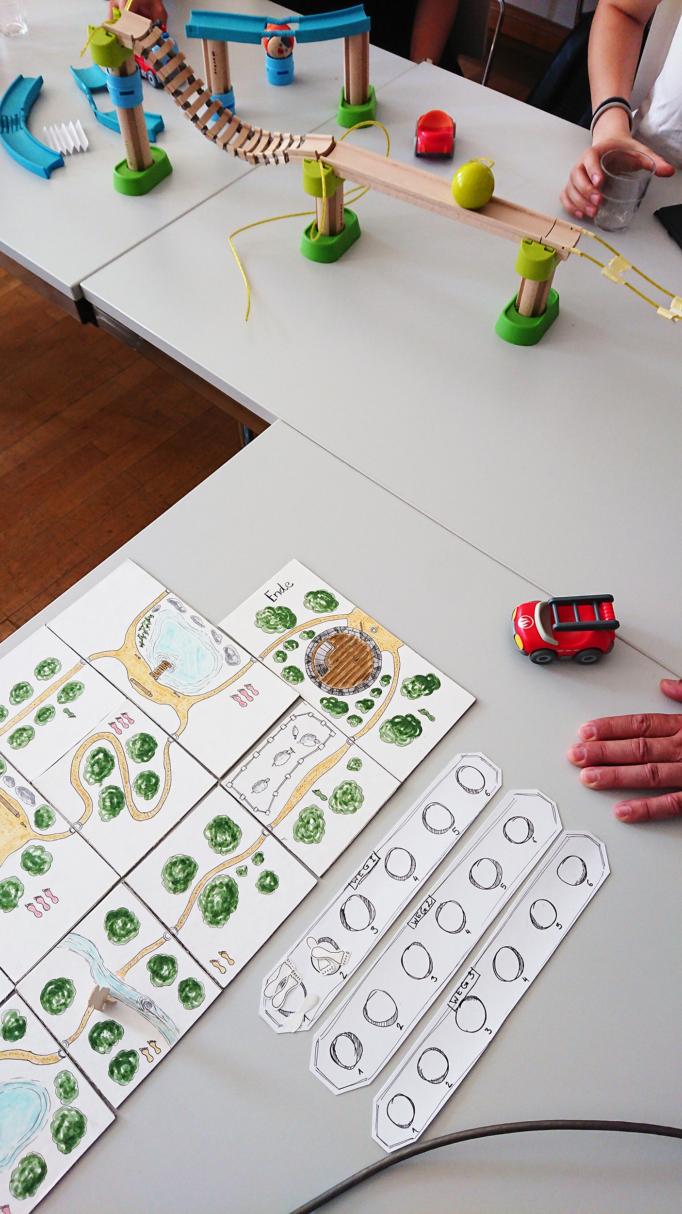 Ein Spielaufbau mit einem Brett, auf dem ein Weg durch Grünflächen, ein rotes Spielzeugauto und Karten abgebildet sind. Im Hintergrund ist, in Anlehnung an die innovativen Designs der Hochschule Coburg, eine Murmelbahn mit Brücken und Rampen auf einem Tisch aufgebaut. Neben dem Brett ist die Hand einer Person zu sehen.