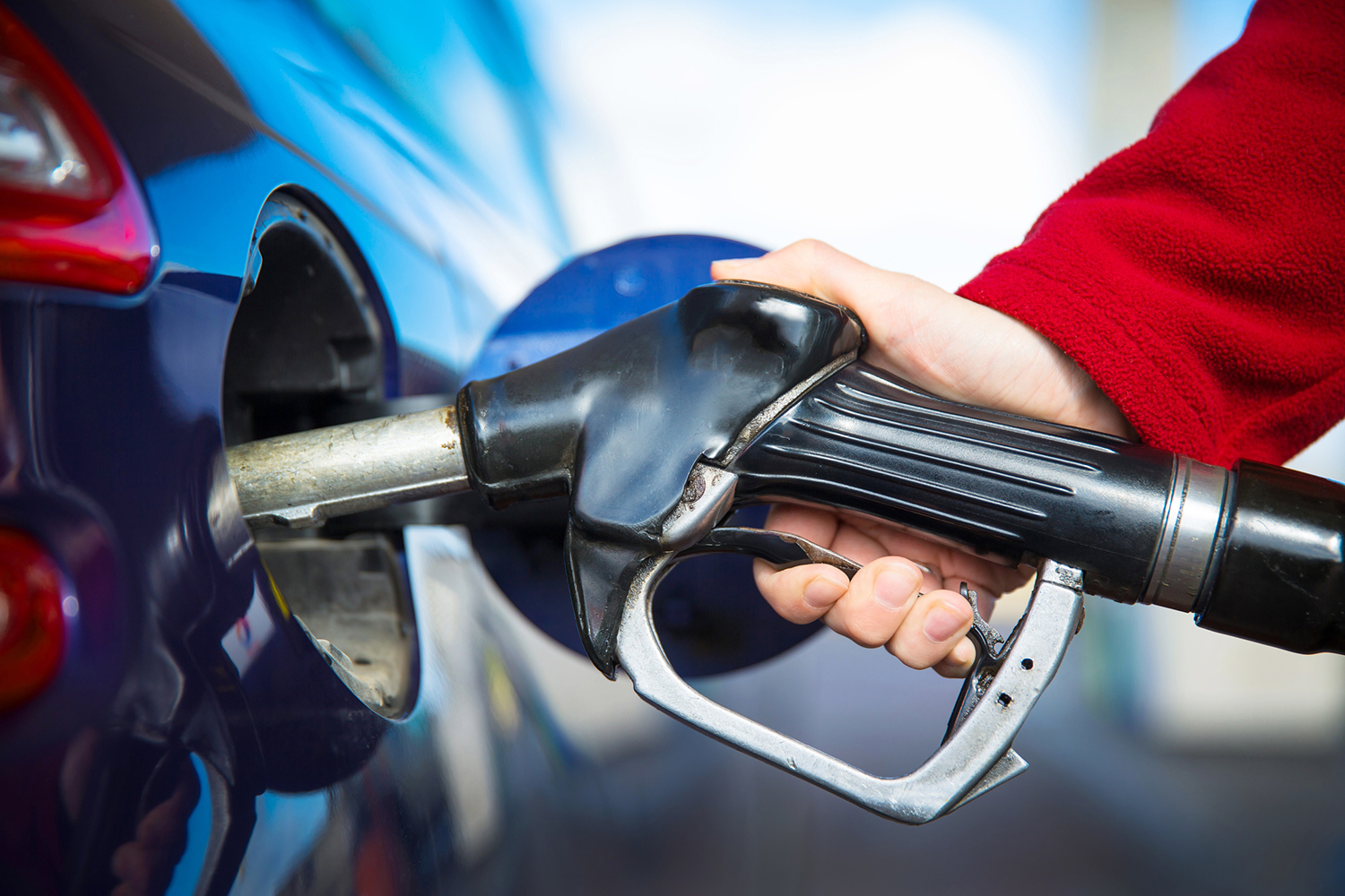 Eine Person in einem roten Ärmel betankt ein Auto an der Tankstelle und führt eine schwarze Zapfpistole in den Tank des Fahrzeugs ein. Der Hintergrund, der an einen geschäftigen Tag in der Nähe der Hochschule Coburg erinnert, ist leicht unscharf und lenkt den Fokus auf die präzise Aktion des Tankens.