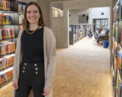 Eine Frau mit langen Haaren, einem schwarzen Hemd und einer beigen Strickjacke steht lächelnd in der Bibliothek der Hochschule Coburg. An den Wänden stehen Regale voller bunter Bücher, im Hintergrund sieht man Menschen, die an Schreibtischen lernen.