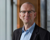 Ein Mann mit Brille, Glatze und einem hellblauen Hemd unter einer dunklen Jacke steht lächelnd im Gebäude der Hochschule Coburg vor einem verschwommenen Hintergrund aus Fenstern und einer Holztafel.