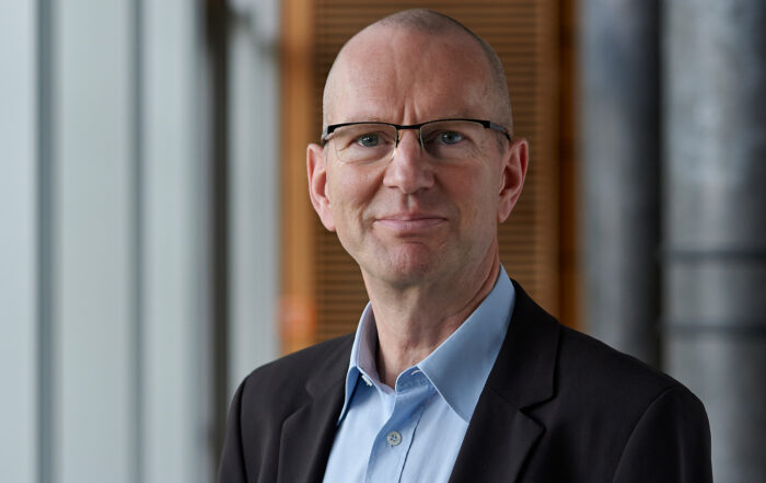 Ein Mann mit Brille, Glatze und einem hellblauen Hemd unter einer dunklen Jacke steht lächelnd im Gebäude der Hochschule Coburg vor einem verschwommenen Hintergrund aus Fenstern und einer Holztafel.