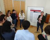 Eine Frau von der Hochschule Coburg hält einen Vortrag vor einer Gruppe, die sich um ein großes Whiteboard mit Postern versammelt hat. Der Raum mit Holzboden und einer Lochwand ist voller aufmerksamer Teilnehmer, von denen einige fleißig Notizen machen.