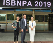 Drei Menschen stehen lächelnd vor einem Gebäudeeingang, über dem ein Schild mit der Aufschrift „ISBNPA 2019“ hängt. Flankiert von den beiden Frauen steht der Mann stolz im Business-Outfit und macht ein unvergessliches Gruppenfoto an der Hochschule Coburg.