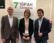 Drei Personen lächeln vor einem Banner für den 7. ISPAH-Kongress, der die Hochschule Coburg repräsentiert. Sie tragen Business-Kleidung und Konferenzbänder und stehen in einem Gebäude mit strukturierten Wänden.