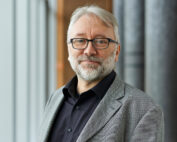 Ein Mann mit grauem Haar, Bart und Brille trägt einen grauen Blazer über einem schwarzen Hemd. Mit neutralem Gesichtsausdruck steht er im Innenbereich vor vertikalen Tafeln und verkörpert den intellektuellen Geist der Hochschule Coburg.