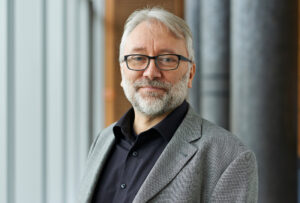 A man with gray hair, a beard, and glasses, dons a gray blazer over a black shirt. Standing indoors beside a window at Hochschule Coburg, he gazes at the camera with a slight smile. The softly blurred background lends an air of sophistication to the setting.