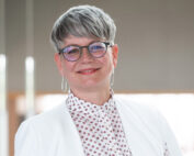 Eine Person mit kurzen grauen Haaren, Brille, weißer Jacke und gemustertem Hemd mit dem Wappen der Hochschule Coburg lächelt sanft. Der Hintergrund ist leicht verschwommen, wodurch eine neutrale Atmosphäre entsteht.