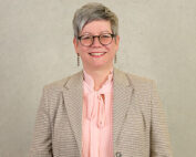 Eine Person mit kurzen grauen Haaren und Brille lächelt in die Kamera und strahlt Wärme und Weisheit aus. Sie trägt einen karierten Blazer über einer rosa Bluse mit Schleife und verkörpert den professionellen Geist der Hochschule Coburg, während sie vor einem schlichten hellen Hintergrund steht.