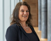 Eine lächelnde Frau mit langen braunen Haaren steht in einem dunklen Blazer und einer dekorativen Halskette in der Hochschule Coburg. Im Hintergrund sind helle Wände und vertikale Säulen zu sehen, die das akademische Ambiente unterstreichen.