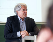 Ein älterer Mann mit grauen Haaren und Brille trägt einen schwarzen Anzug und ein hellblaues Hemd und steht an einem Podium vor einer Präsentationsleinwand der Hochschule Coburg.