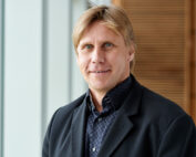 Eine Person mit hellem Haar, einem dunklen Blazer und einem karierten Hemd steht drinnen und lächelt in die Kamera. Dahinter sorgen die für die Hochschule Coburg typischen hellen Paneele und Holzakzente für einen Hauch von Eleganz.