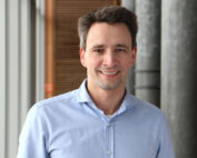 Ein Mann mit kurzen dunklen Haaren und einem hellblauen Hemd lächelt in die Kamera. Er steht in einem Gebäude der Hochschule Coburg, im Hintergrund sind vertikale Tafeln und eine Säule zu sehen.