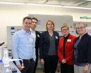 Fünf Personen stehen zusammen in einem Labor an der Hochschule Coburg und lächeln in die Kamera. Sie sind in legerer Business-Kleidung gekleidet und tragen Ausweise, umgeben von einer Reihe von Laborgeräten und Maschinen.