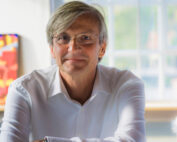 Eine Person mit grauem Haar und Brille lächelt und trägt ein weißes Hemd. Im Innenbereich der Hochschule Coburg bestimmen lebendige Kunstwerke und ein helles Fenster die Szenerie.