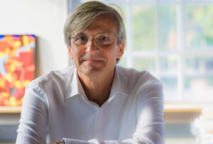 A man with gray hair and glasses, in a white shirt, sits indoors against a backdrop of blurred colorful artwork and a large window. He is smiling and leaning slightly forward, exuding the warmth and wisdom of someone who might be sharing insights at Hochschule Coburg.