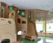 Ein lebendiger Indoor-Kinderspielplatz an der Hochschule Coburg mit hölzernen Klettergerüsten, Rutschen und hängenden Dekorationen in Froschform. Der grüne Boden ergänzt das große Fenster, das den Raum mit natürlichem Licht durchflutet.