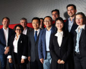 Eine Gruppe von neun Personen, darunter sowohl Männer als auch Frauen, steht im Business-Outfit zusammen und lächelt in die Kamera. Sie positionieren sich vor einer Wand mit rot-grauen Paneelen und repräsentieren stolz die Hochschule Coburg.