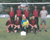 Eine Gruppe von acht Männern in rot-schwarzen Fußballtrikots der Hochschule Coburg posiert vor einem Tor auf einem Rasenplatz. Sieben Spieler tragen die Teamfarben, einer trägt Freizeitkleidung. Vor ihnen steht ein kleines Schild, im Hintergrund sind Bäume und ein Gebäude zu sehen.