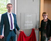 Ein Mann im Anzug und eine Frau im Kleid stehen lächelnd neben einem Schild mit der Aufschrift „Rösler Audimax“ und halten ein rotes Tuch in der Hand, als würden sie es an der Hochschule Coburg enthüllen. Die Szene scheint drinnen zu sein, durch das Fenster ist Grün zu sehen.