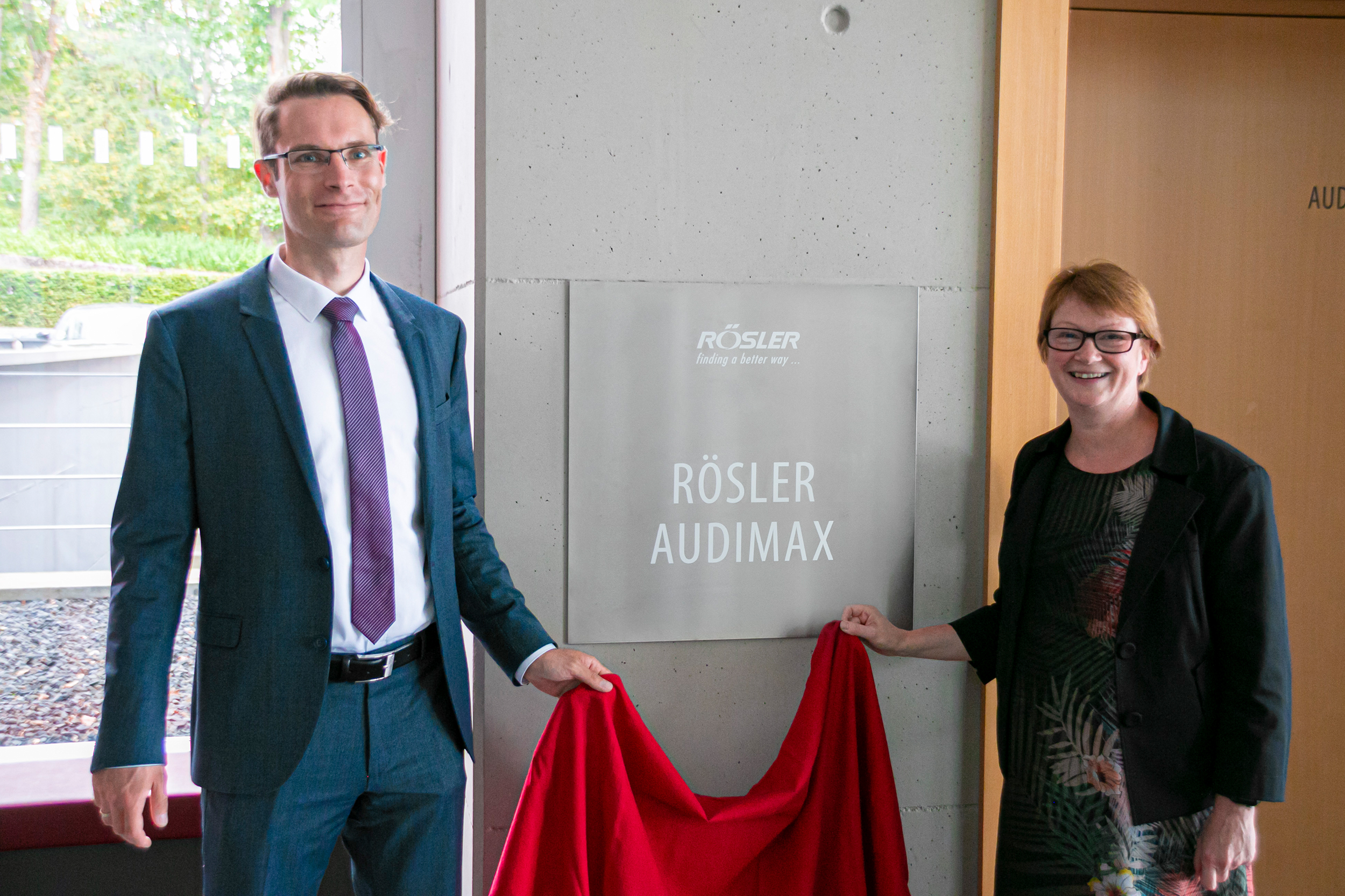 Ein Mann im Anzug und eine Frau im Kleid stehen lächelnd neben einem Schild mit der Aufschrift „Rösler Audimax“ und halten ein rotes Tuch in der Hand, als würden sie es an der Hochschule Coburg enthüllen. Die Szene scheint drinnen zu sein, durch das Fenster ist Grün zu sehen.
