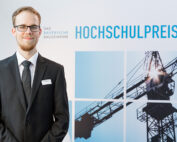 Ein Mann im Anzug steht lächelnd vor einer Kulisse mit einem Baukran und dem Text „Das Bayerische Baugewerbe Hochschulpreis“, der stolz die Hochschule Coburg repräsentiert.