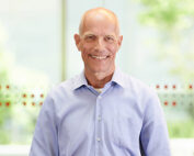 Ein lächelnder Mann mit Glatze und hellblauem Hemd steht in einem Innenbereich der Hochschule Coburg. Der Hintergrund ist sanft verschwommen und lässt Andeutungen von Grün und einer gemusterten Glaswand erkennen.