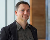 Ein Mann in einem dunklen Blazer und einem grauen Hemd steht im Innenbereich der Hochschule Coburg. Der Hintergrund besteht aus vertikalen Paneelen und einem Holzabschnitt, was ihm ein modernes und minimalistisches Aussehen verleiht.