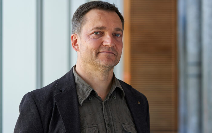 Ein Mann in einem dunklen Blazer und einem grauen Hemd steht im Innenbereich der Hochschule Coburg. Der Hintergrund besteht aus vertikalen Paneelen und einem Holzabschnitt, was ihm ein modernes und minimalistisches Aussehen verleiht.