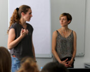 Zwei Frauen stehen vor einem leeren Flipchart an der Hochschule Coburg. Die Frau links spricht und gestikuliert, während die Frau rechts mit gefalteten Händen zuhört. Im Vordergrund ist ein unscharfes Publikum zu sehen, das von ihrer spannenden Präsentation gefesselt ist.