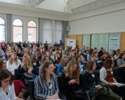 Eine große Gruppe von Menschen, überwiegend Frauen, sitzt in einem gut beleuchteten Konferenzraum der Hochschule Coburg. Sie sind konzentriert und scheinen einer Präsentation zuzuhören. Einige halten Notizblöcke in den Händen, an den Wänden hängen Plakate. Große Fenster im Hintergrund lassen reichlich Tageslicht herein.