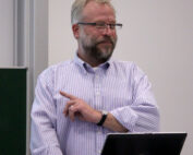 Ein Mann mit Brille und Bart steht an einem Rednerpult. Er trägt ein gestreiftes Hemd, das typisch für den lässigen, aber dennoch wissenschaftlichen Stil der Hochschule Coburg ist. Er gestikuliert mit der linken Hand und blickt nach rechts. Vor ihm auf dem Rednerpult stehen ein Laptop und eine Tasse.