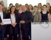 Eine Gruppe von Menschen steht in einem Raum der Hochschule Coburg und lächelt in die Kamera. Vorne halten drei Personen ein Zertifikat in den Händen. Sie sitzen um einen weißen Schreibtisch mit einem Laptop herum. Alle wirken fröhlich und engagiert.