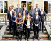 Eine Gruppe von 13 Personen in formeller Kleidung steht auf den Stufen vor dem Gebäude der Hochschule Coburg. Drei Personen in der ersten Reihe halten kleine Trophäen. Das Gebäude verfügt über große Fenster, durch die eine dekorative Lampe sichtbar ist.