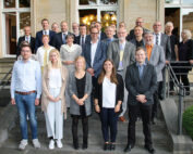 Ein Gruppenfoto zeigt Menschen, die auf den Stufen vor einem Gebäude stehen, möglicherweise der Hochschule Coburg. Die Männer und Frauen in formeller und halbformeller Kleidung posieren lächelnd zusammen, was auf eine Versammlung oder Veranstaltung im Freien schließen lässt.