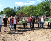 Eine Gruppe von neun Personen steht mit drei Pferden auf einem Sandgelände, vermutlich auf einer Bildungsexkursion der Hochschule Coburg. Im Hintergrund sind Bäume und ein kleines Gebäude zu sehen, unter einem klaren und sonnigen Himmel.