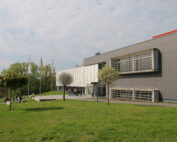 Das moderne Gebäude der Hochschule Coburg zeichnet sich durch eine elegante graue Fassade, große Fenster und einen einzigartigen weißen Bereich aus. Flaggen säumen den Gehweg, während sich Menschen in der Nähe des Eingangs versammeln, während eine Rasenfläche mit Bäumen das Gebäude unter einem teilweise bewölkten Himmel umgibt.
