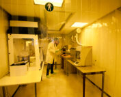 Im gelb beleuchteten Reinraum der Hochschule Coburg navigiert eine Person in weißem Laborkittel und grünen Überschuhen durch verschiedene Laborgeräte. Transparente Kunststoffvorhänge rahmen den Hightech-Arbeitsbereich ein und sorgen für Präzision und Sicherheit.