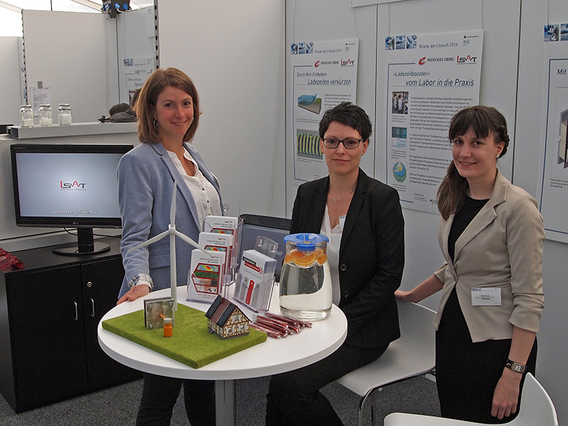 Am Messestand präsentieren drei Frauen der Hochschule Coburg auf einer Miniaturlandschaft ein Windrad und Solarpanele. Plakate im Hintergrund liefern Details, links flimmert ein Fernseher und bietet ein spannendes visuelles Erlebnis.