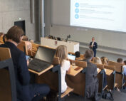 In einem modernen Hörsaal der Hochschule Coburg mit Sitzreihen hält ein Dozent einen Vortrag, während die Teilnehmer, von denen einige Laptops tragen, aufmerksam zuhören. Vorne zeigt ein großer Bildschirm Informationen vor dem Hintergrund des eleganten Beton- und Holzdesigns des Saals.