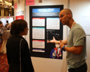 Zwei Personen unterhalten sich vor einem Poster mit dem Titel „Temporale Trends“ in der Ausstellung der Hochschule Coburg. Das Poster enthält Text, Grafiken und Abbildungen. In der Umgebung herrscht reges Treiben mit Konferenzteilnehmern und im Hintergrund sind verschiedene interessante Darstellungen zu sehen.